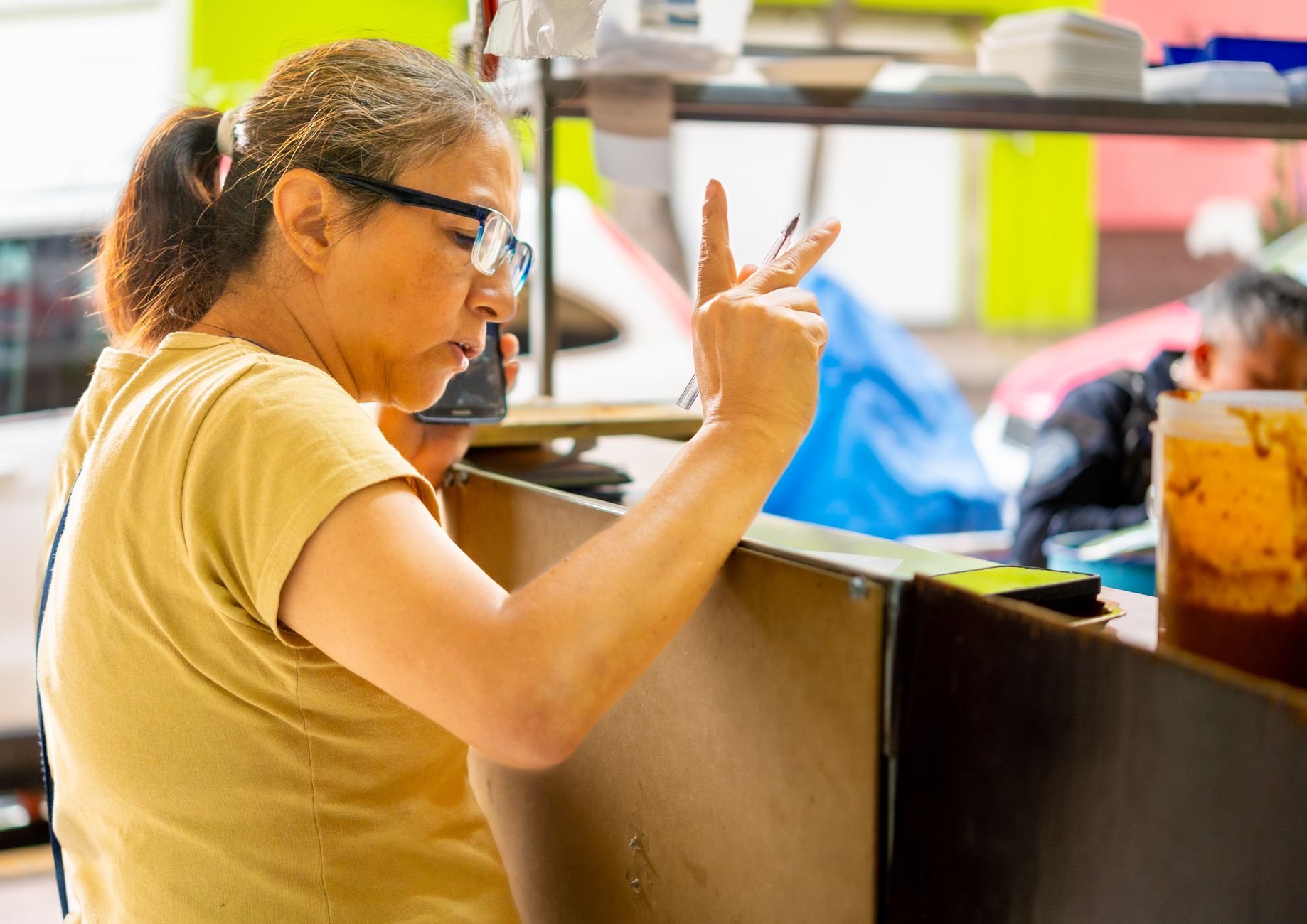 A female vendor takes an order using a smartphone at a lively outdoor food stall.